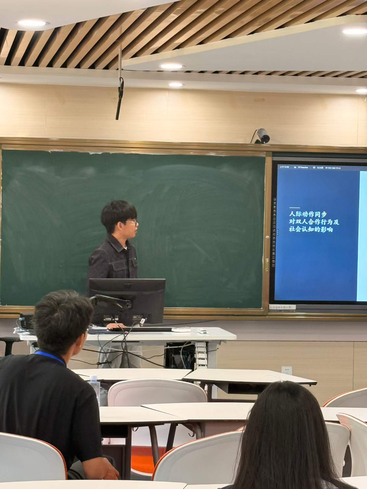 A speaker beside a slide titled The Effects of Interpersonal Movement Synchrony on Dyadic Cooperation and Social Cognition